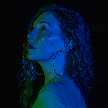 A profile image of Angela Dix who has long hair, and is wearing large hoop earings. The image is dark, they are looking away into the distance, and their face is illuminated by blue and green neon light.