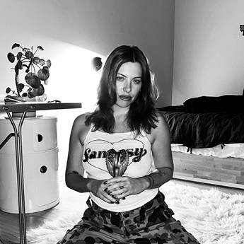 A profile picture of Micha Reads Stars. The image is black and white and shows Micha in a room, kneeling on the floor. They are holding an ornament in their hands, and are wearing a white shirt and patterned pants.