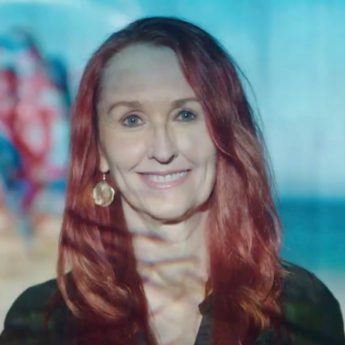 A headshot photograph of Jill Bennett who is smiling and has long red hair. An image is being projected across her face. She is wearing gold earrings and a black top.