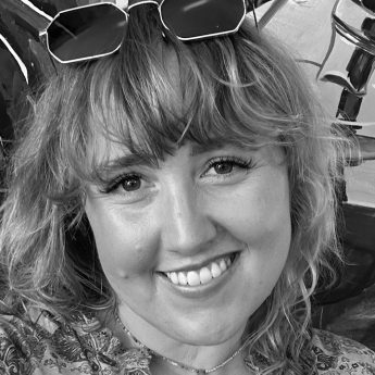 A black and white photograph of a woman with blonde hair smiling at the camera. She has a patterned shirt on and sunglasses on her head.