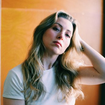 A photograph of Ash King in a white t-shirt with long blonde hair. She is leaning her head into her hand and is standing in front of a bright orange background.