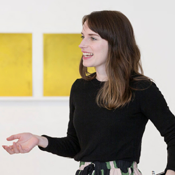 A person with long brown hair smiling with their body angled towards the left. They are standing in front of a series of yellow artworks. They are wearing a black long-sleeved shirt and a floral skirl.