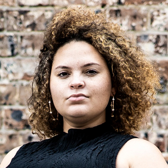 A headshot image of Kaleigh Giles. Kaleigh has short curly brown hair and is wearing a black turtle neck singlet. They have hoop earrings on and are looking into the camera with a confident expression on their face. They are standing in front of a brick wall.