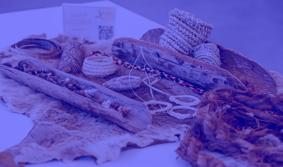 A series of woven Aboriginal crafts sit on a possum rug on top of a table. There is a purple filter over the image.