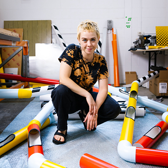 A photograph of artist Nadia Odlum in their studio. Nadia is wearing a tie-dye black and orange t-shirt and black pants. They have short, cropped blonde hair. They are crouching down amongst a series of bright coloured sculptures that resemble pipes.